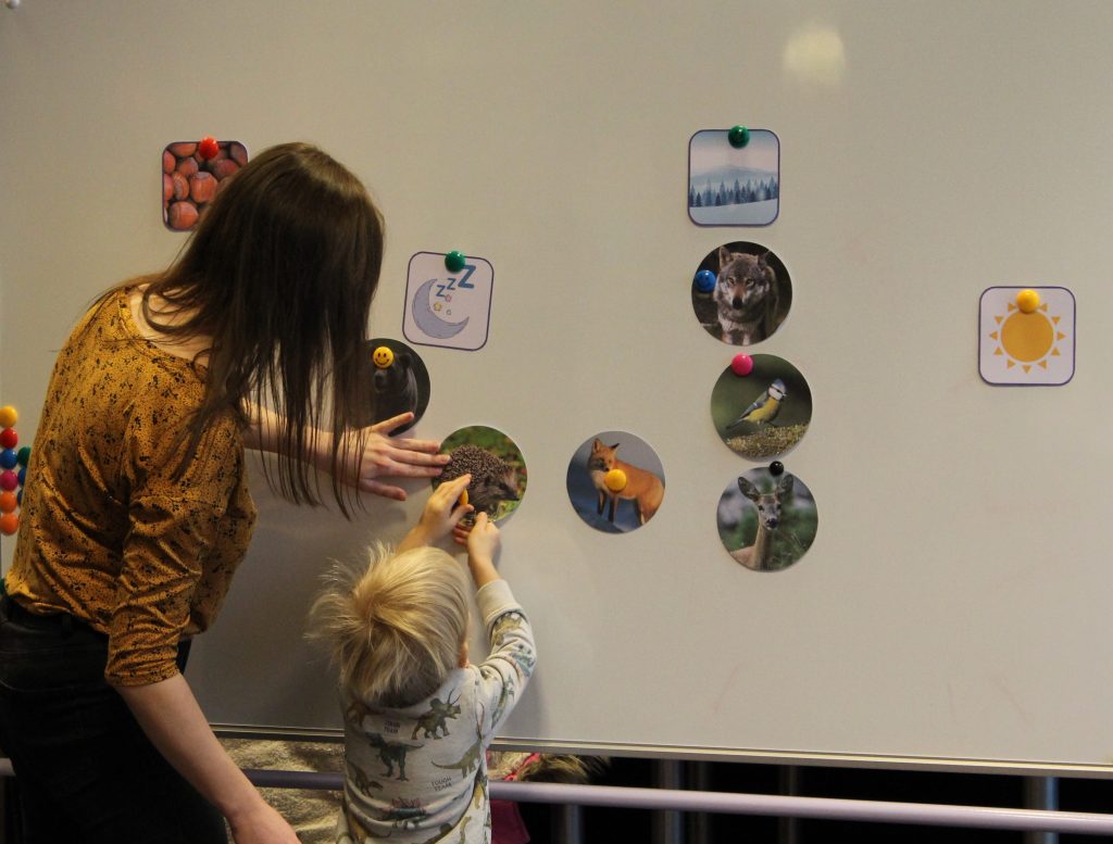 Na zdjęciu bibliotekarka pomaga chłopcu zawiesić zdjęcie na tablicy magnetycznej; na tablicy jest kilka zdjęć zwierząt i symbole oznaczające sposób spędzania zimy (sen zimowy, robienie zapasów, aktywne spędzanie zimy, odlot do ciepłych krajów).