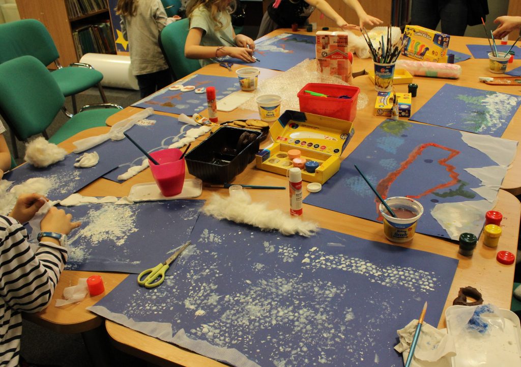 Na zdjęciu stół podczas wykonywania pracy plastycznej przez dzieci; na blacie kawałki brystolu, farby, kubki z wodą, wata, nożyczki, kleje itp.
