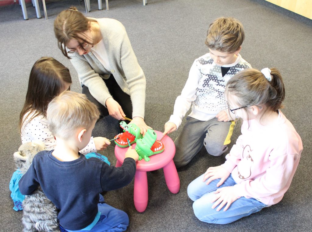 Na zdjęciu bibliotekarka i czworo dzieci. Dzieci ołówkami naciskają zęby dwóch zabawkowych krokodyli stojących na taborecie.