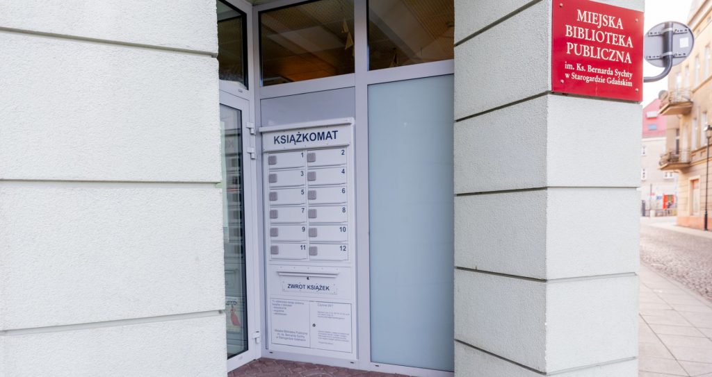 Wejście do Miejskiej Biblioteki Publicznej w Starogardzie Gdańskim z widocznym białym Książkomatem wbudowanym w przeszklone drzwi. Na urządzeniu znajduje się 12 ponumerowanych skrytek oraz wrzutnia z napisem "Zwrot Książek". Obok, na jasnej elewacji budynku, wisi czerwona tablica informacyjna z godłem Polski i nazwą biblioteki. 